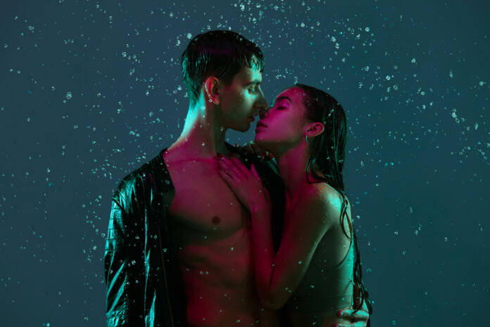 Tender kisses. Young couple, man and woman hugging, kissing under the rain over blue background in neon light. Concept of human emotions and feelings, love, passion, tenderness