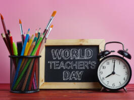 World Teacher's Day Text. Blackboard, Alarm Clock and School Stationary in Basket on Wooden Table Pink Background