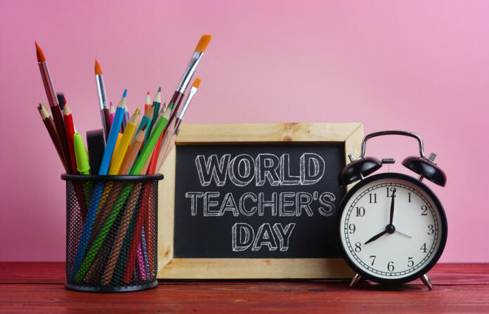 World Teacher's Day Text. Blackboard, Alarm Clock and School Stationary in Basket on Wooden Table Pink Background