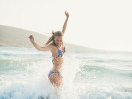 Young beautiful blond woman tourist in swimsuit jumping and having fun in wavy waters of Mediterranean sea in Prasonisi cape, Rhodes, Greek summer resort. Travel, vacation, freedom, happiness