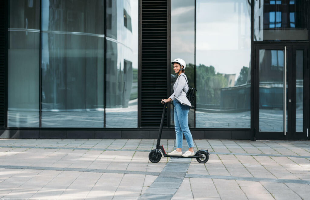 Young businesswoman wearing cycling helmet riding push scooter on footpath