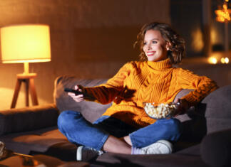 Ker nekateri še vedno nis(m)o pogledali serije The Game of Thrones in/ali The Witcher young cheerful woman eating popcorn and watches movie on cable TV while switching channels with the remote control at home in evening alone