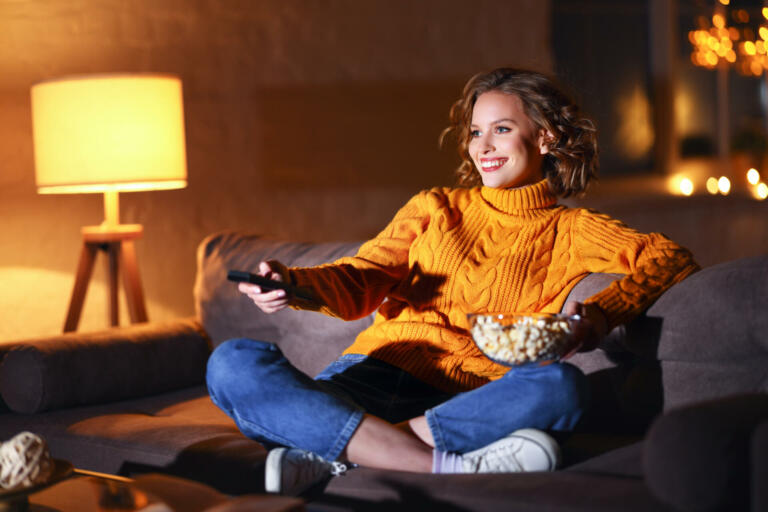 young  cheerful woman eating popcorn and watches  movie on  cable TV while switching channels with the remote control at home in evening  alone