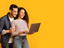 Young middle eastern couple using laptop together while standing over yellow background in studio, cheerful arab spouses shopping online or browsing new website on computer, panorama with copy space