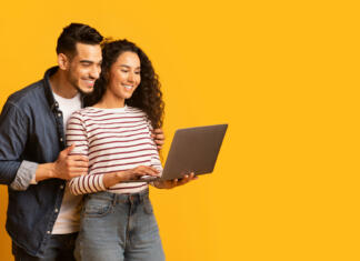 Na kaj moramo biti pozorni pri nakupu prenosnika? Young middle eastern couple using laptop together while standing over yellow background in studio, cheerful arab spouses shopping online or browsing new website on computer, panorama with copy space