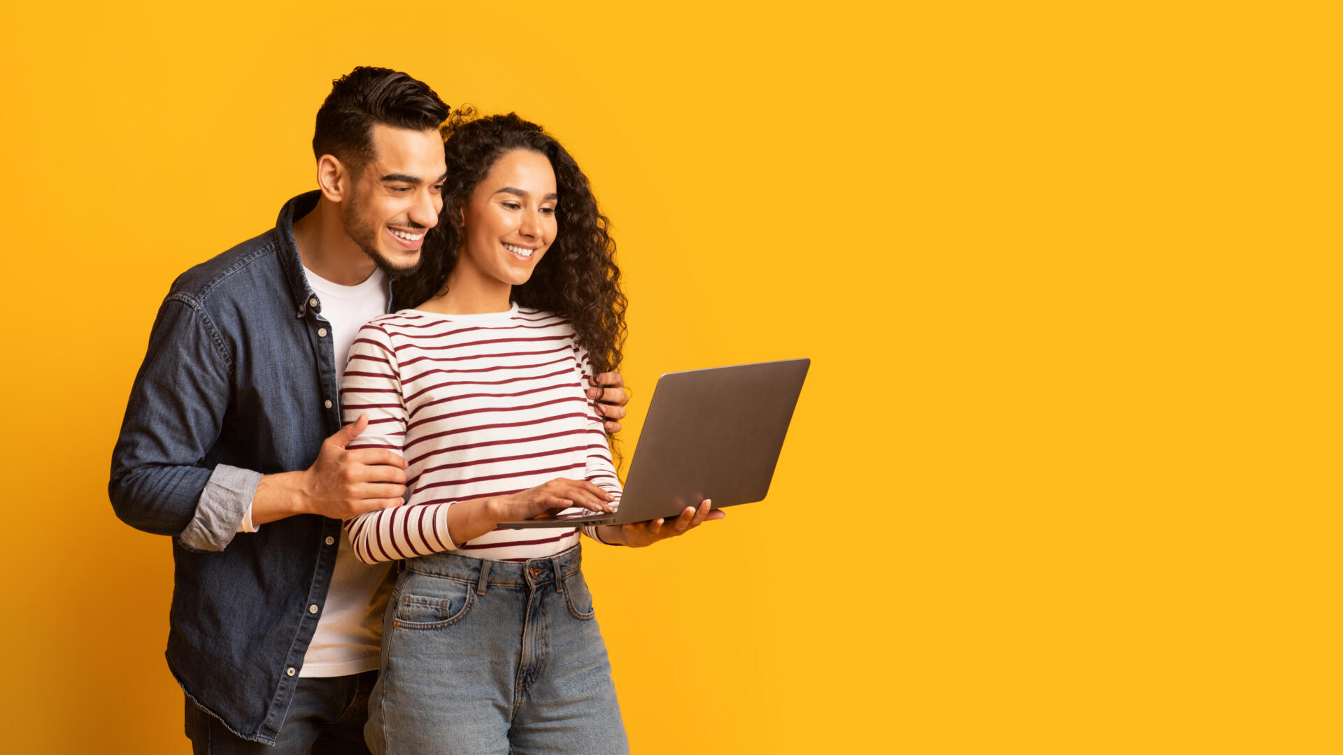 Young middle eastern couple using laptop together while standing over yellow background in studio, cheerful arab spouses shopping online or browsing new website on computer, panorama with copy space