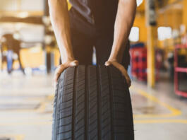 A handsome young mechanic in white gloved uniform is putting his hand on a new tire and checking and checking the condition of the tire while doing car service.
