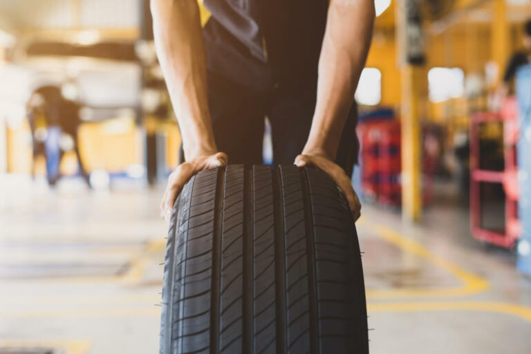 A handsome young mechanic in white gloved uniform is putting his hand on a new tire and checking and checking the condition of the tire while doing car service.