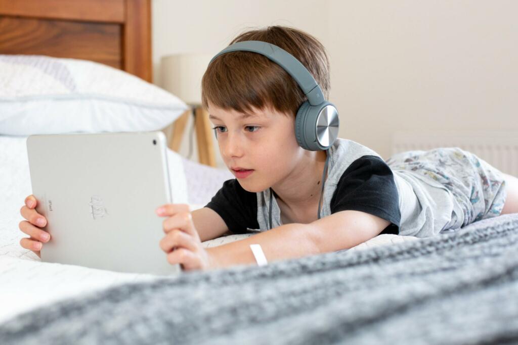 Deček s slušalkami gleda v tablični računalnik in leži na postelji