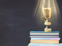 Golden trophy on pile of books, against blackboard, with sun rays over trophy; learning/achievement concept
