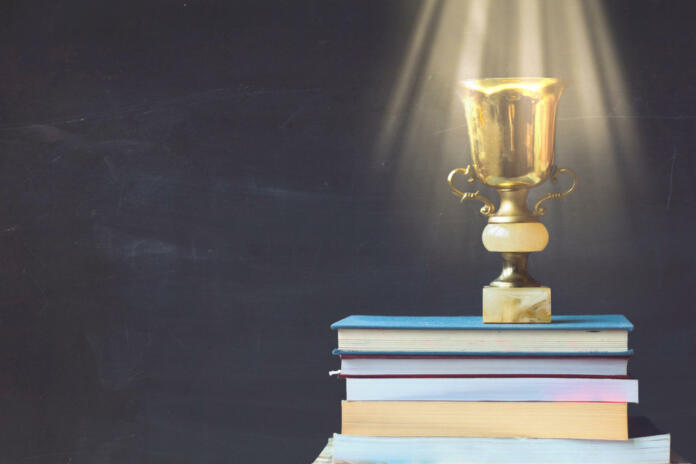 Golden trophy on pile of books, against blackboard, with sun rays over trophy; learning/achievement concept