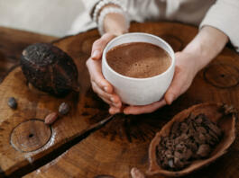 Ženska v roki drži skodelico vroče čokolade, poleg nje je na lesenem pultu kakav