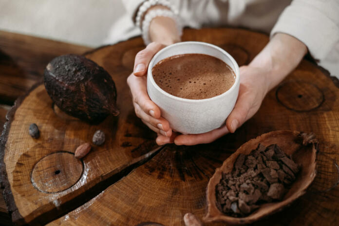 Ženska v roki drži skodelico vroče čokolade, poleg nje je na lesenem pultu kakav