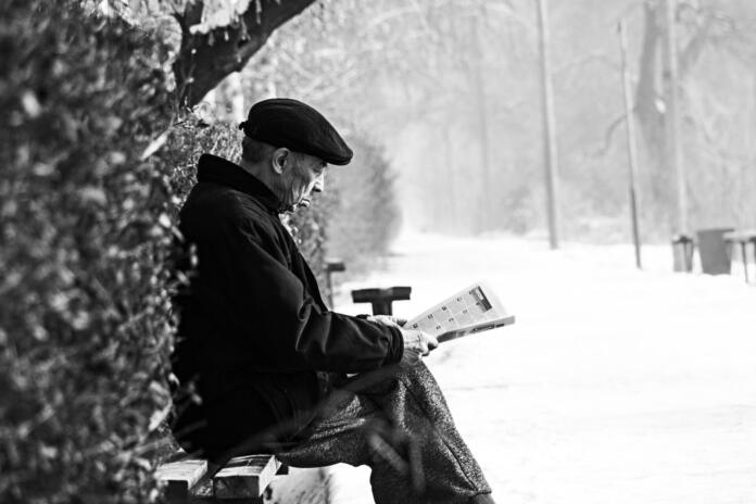 Star moški med sneženjem sedi na klopi in bere knjigo