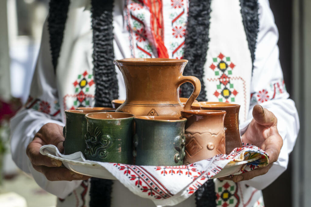 Moški v izvezenih tradiciionalnih oblačilih nosi pladenj z glinenimi lončki in vrčem