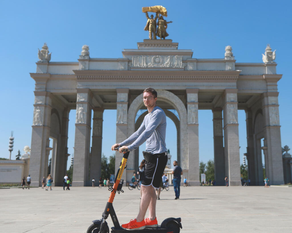 Moscow, Russia - 07.12.2021: Young man in summer clothes on an electric scooter at the entrance to Exhibition of Achievements of National Economy (Russian: aka VDNKh or VDNH an amusement park)