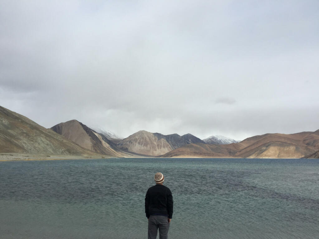 Person standing facing body of water with hills at distance