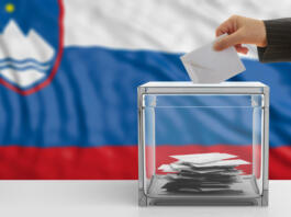 Oseba meče volilni listek v škatlo pred slovensko zastavo