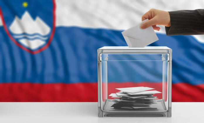 Oseba meče volilni listek v škatlo pred slovensko zastavo