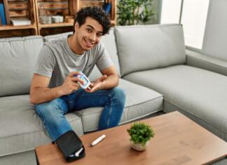 Kajh je diabetes ali sladkorna bolezen? Young hispanic diabetic man measuring glucose sitting on the sofa at home.