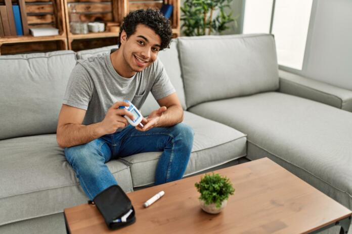 Young hispanic diabetic man measuring glucose sitting on the sofa at home.