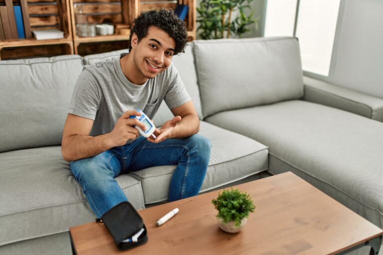 Young hispanic diabetic man measuring glucose sitting on the sofa at home.