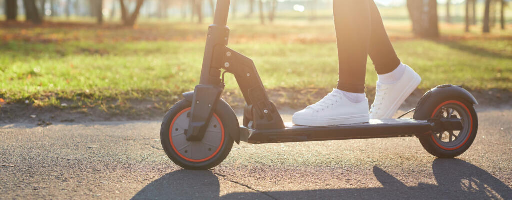 Young woman discover city and park at sunset with electric scooter or e-scooter. Female Legs in sports sneakers stand on electric scooter. Girl riding on Ecological and urban transport.