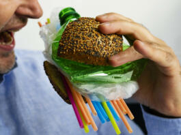 A man eats a burger made from a tasty bun with sesame and poppyseed filled with different types of disposable plastic. The concept of disposable plastic as a danger for ecology and human health.