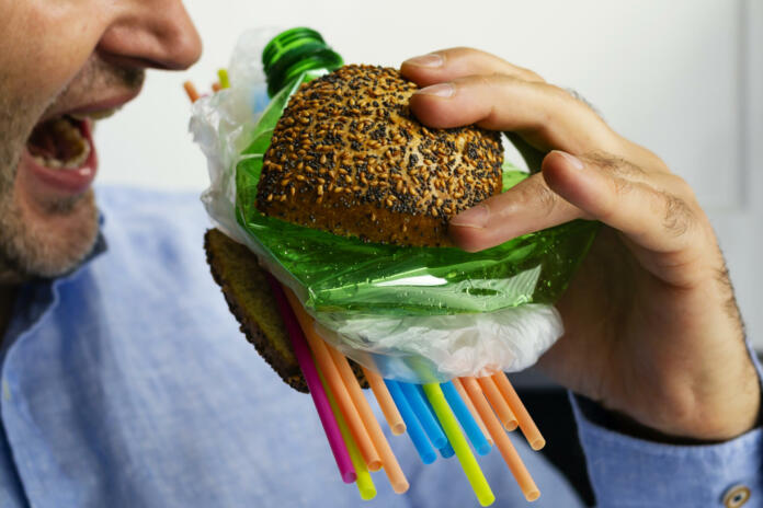 A man eats a burger made from a tasty bun with sesame and poppyseed filled with different types of disposable plastic. The concept of disposable plastic as a danger for ecology and human health.