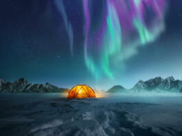 A tent pitched up in snow at night with the northern lights flickering in the sky above. Aurora Borealis and travelling. Photo composite.