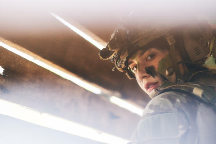 Close up view through hole of wood wall of beautiful soldier woman with combat suit look to the camera.