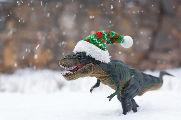 Realistic toy Tyrannosaurus Rex in a small Christmas hat in the snow during a snowfall on the background of a stone wall