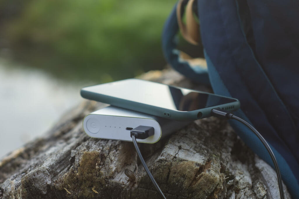 Smartphone is charged using a portable charger. Power Bank charges the phone outdoors with a backpack for tourism in the background of nature and the river