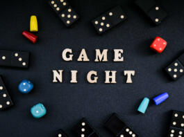 Text GAME NIGHT spelled out in wooden letter. Surrounded by dice, dominoes other game pieces on black background. Table games. Stay home activity for kids family.