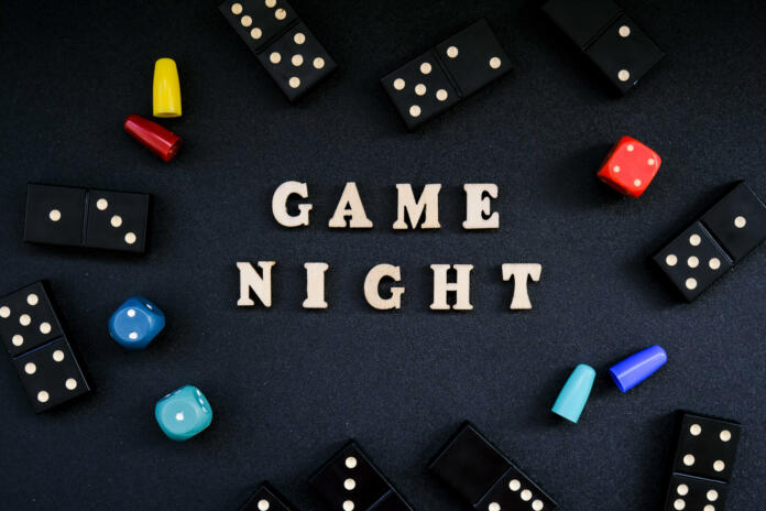 Text GAME NIGHT spelled out in wooden letter. Surrounded by dice, dominoes other game pieces on black background. Table games. Stay home activity for kids family.