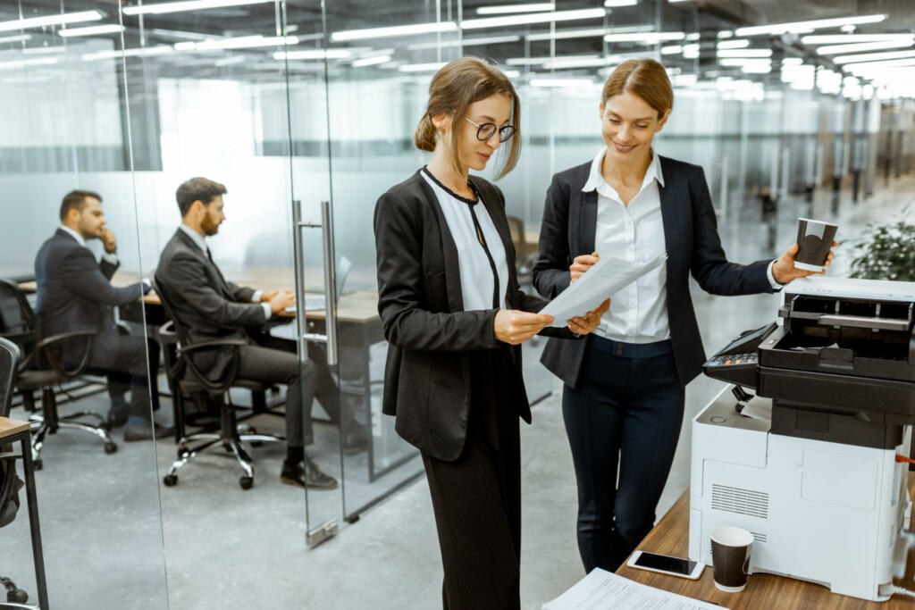 Two business ladies talking near the copier during a coffee break in the hallway of the big corporation