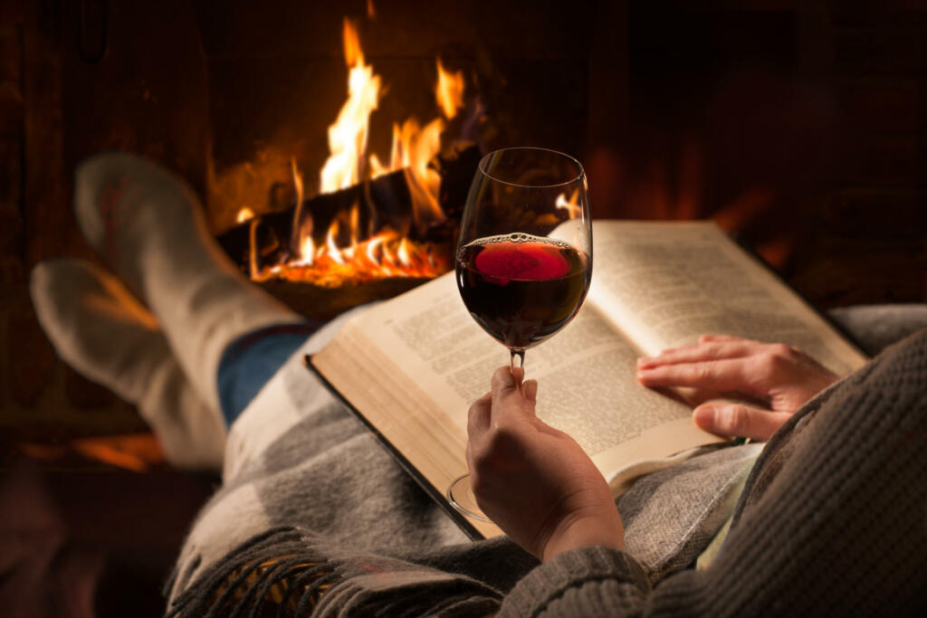 Woman resting with glass of red wine and book near fireplace
