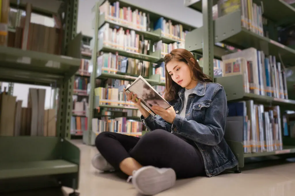 Asian female student sitting on floor in the library, Open and learning textbook from bookshelf in the International College/University Library.