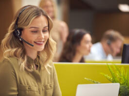 Businesswoman In Cubicle Wearing Headset Talking To Caller In Busy Customer Services Centre