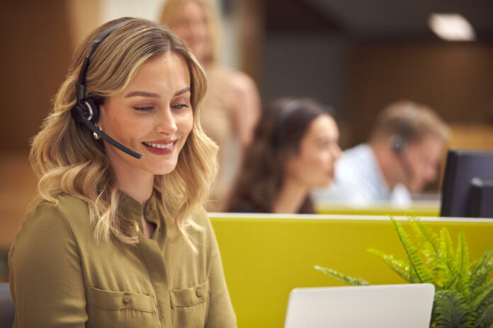 Businesswoman In Cubicle Wearing Headset Talking To Caller In Busy Customer Services Centre