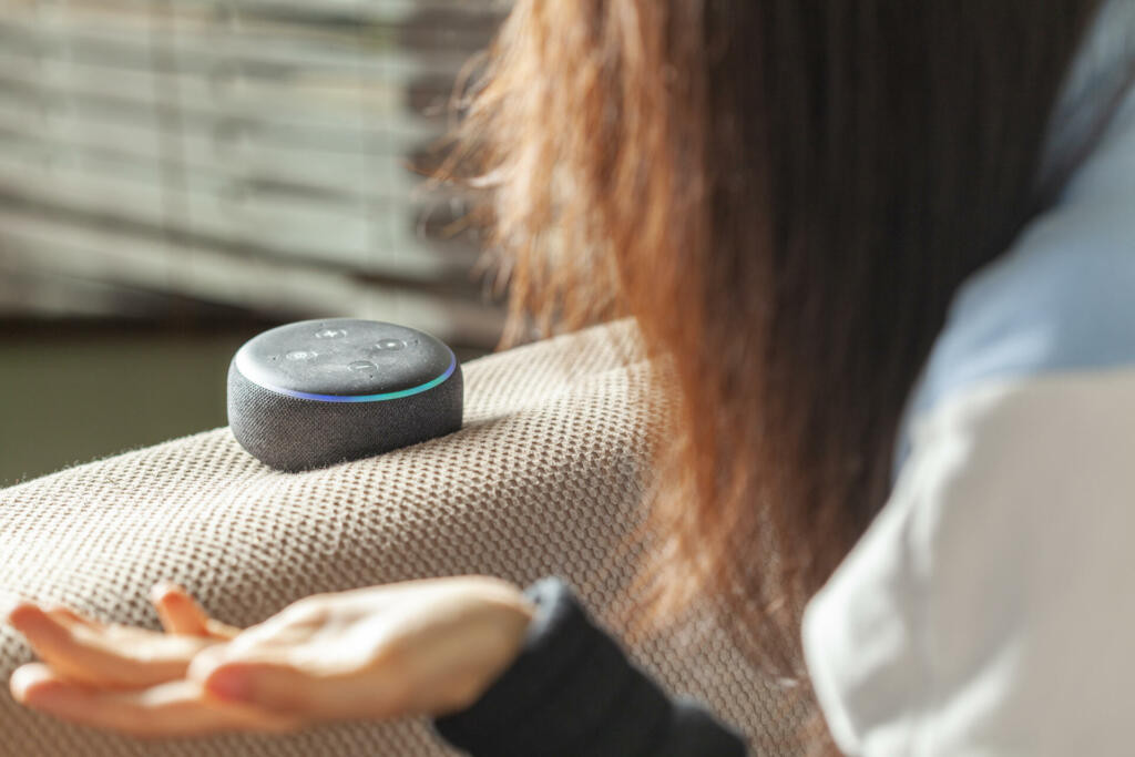 Clarksburg, MD, USA 03-01-2021: A caucasian woman sitting on an armchair with casual clothes in her house is asking a question to Amazon Echo Dot, voice activated speaker device.