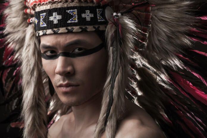Naked indian strong man with traditional native american make up and headdress looking at the camera. Close up desaturated studio shot