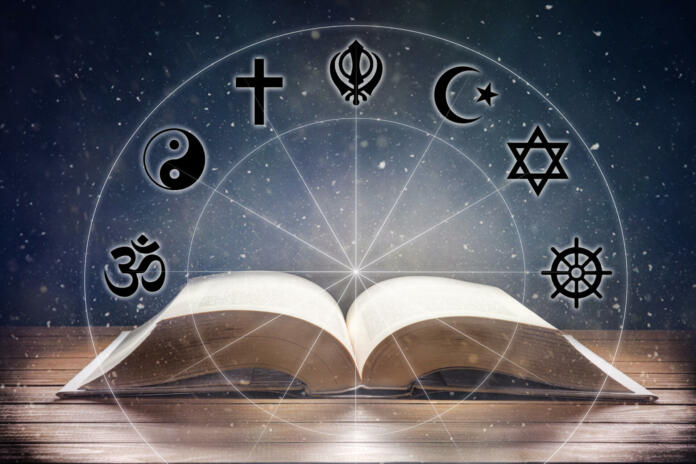 Open book on wooden table with religious symbols and universe background. Concept of the study of religions. Front view.