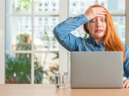 Redhead woman using computer laptop at home stressed with hand on head, shocked with shame and surprise face, angry and frustrated. Fear and upset for mistake.