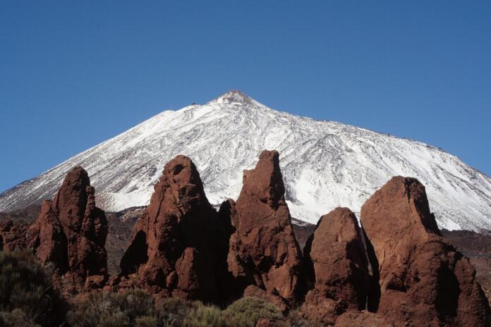 Zasnežen vrh in špičaste skale rdečkastorjave barve v ospredju