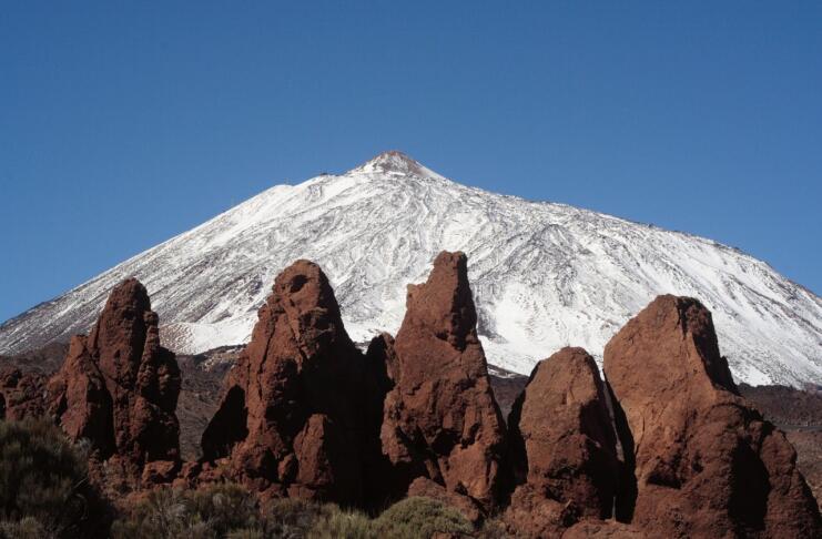 Zasnežen vrh in špičaste skale rdečkastorjave barve v ospredju