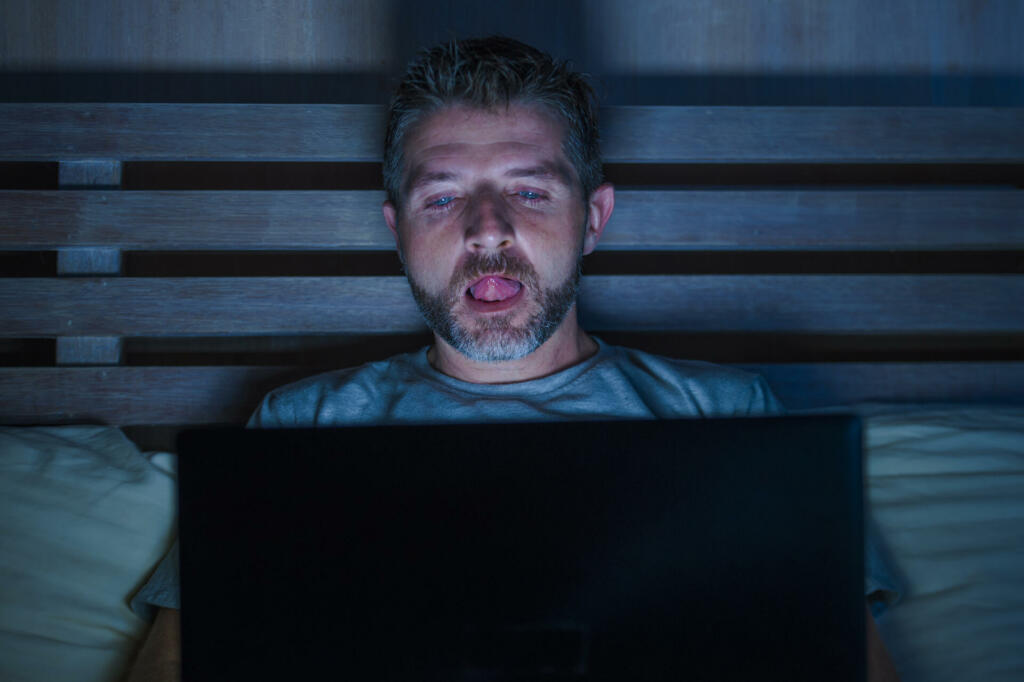 young aroused man alone in bed playing cybersex using laptop computer watching porn sex movie late at night with lascivious pervert face expression in internet pornographic sexual content