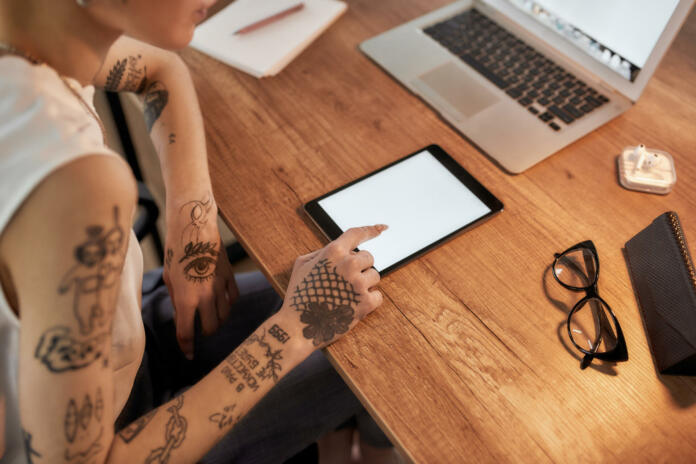 Young tattooed woman with short haircut using digital tablet while sitting at her workplace. Business woman at work. Cropped view. Workplace. Business concept