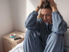 Anxious young woman wearing pajamas, sitting on bed in the morning, holding head in hands, crying
