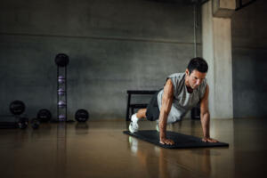 Athletic man doing pushups exercise in sportswear, trains doing plank pose at dark gym floor, looking down, indoors. Dramatic dark shadow background. Copy-space.